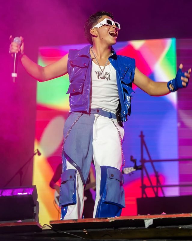 Tony, de Picus, canta con los brazos abiertos, lentes blancos y chaleco azul