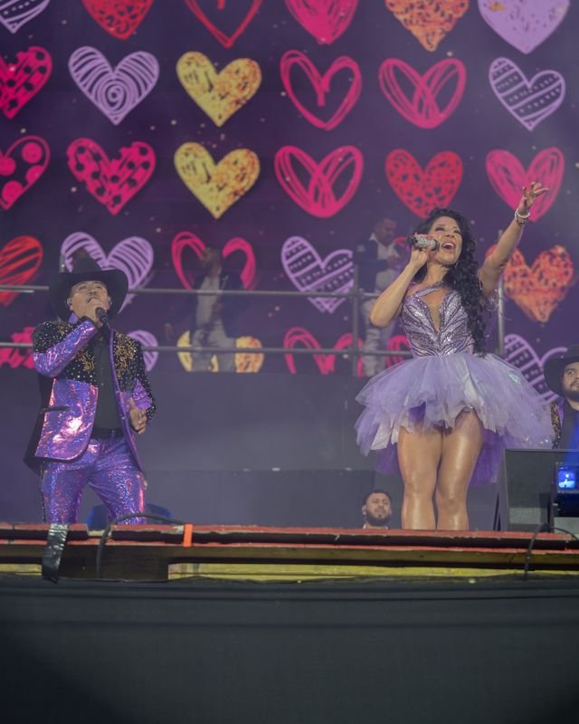 Una cantante de vestido lila con falda de tul levanta el brazo; a su lado canta un hombre de traje morado brillante y sombrero, frente a una pantalla de corazones