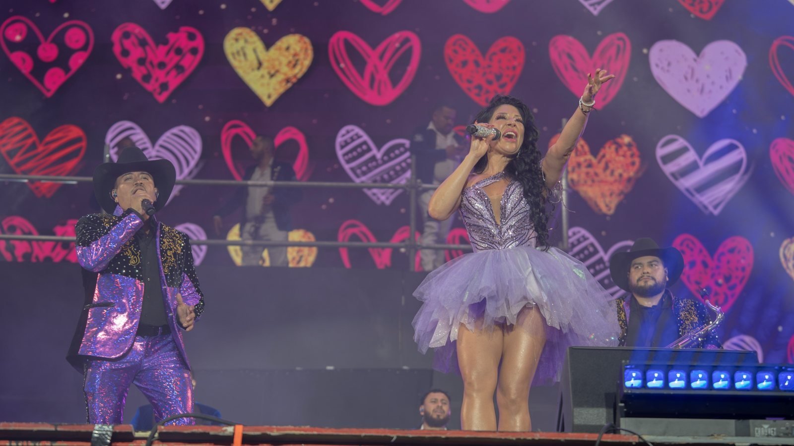 Una de las hermanas Terrazas canta con el brazo en alto frente a una pantalla llena de corazones; a su lado, un cantante de traje morado y sombrero