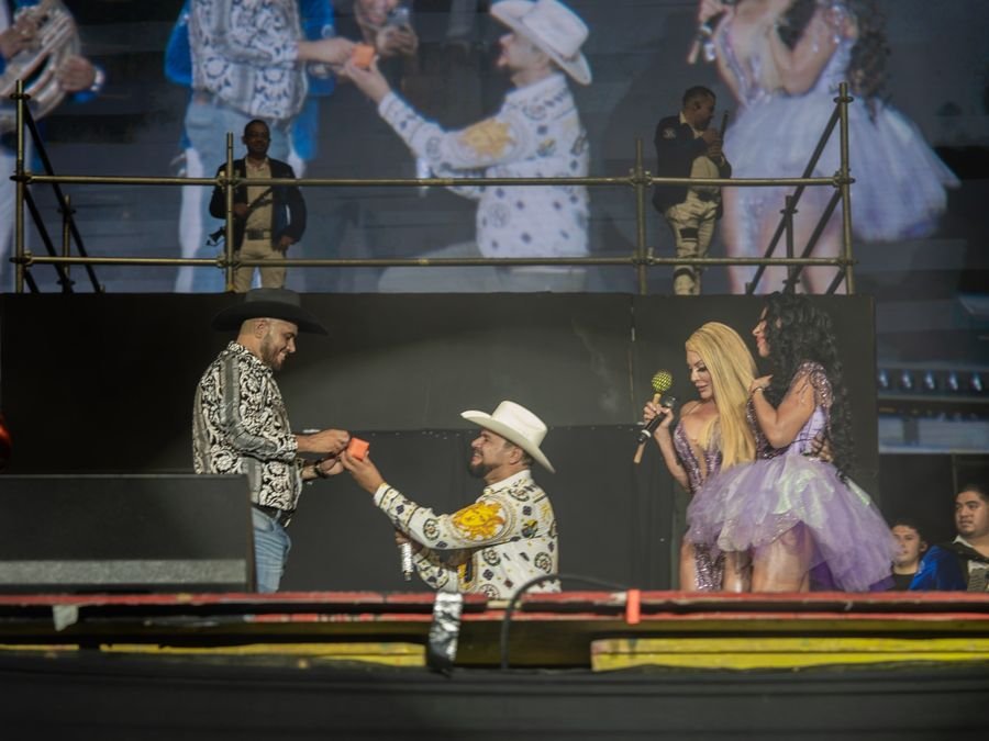 Un asistente de sombrero blanco, hincado en el escenario, entrega una cajita a su pareja mientras las dos cantantes miran abrazadas; la pantalla proyecta el momento