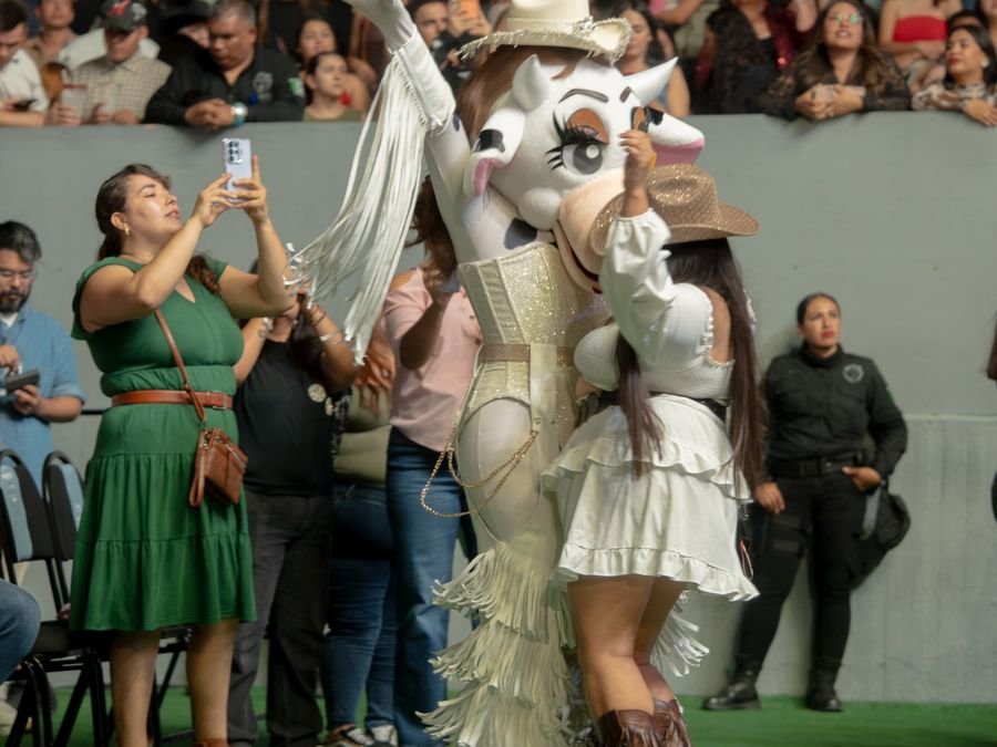 Una botarga de vaquita con sombrero y flecos blancos baila con una asistente mientras el público graba con sus celulares