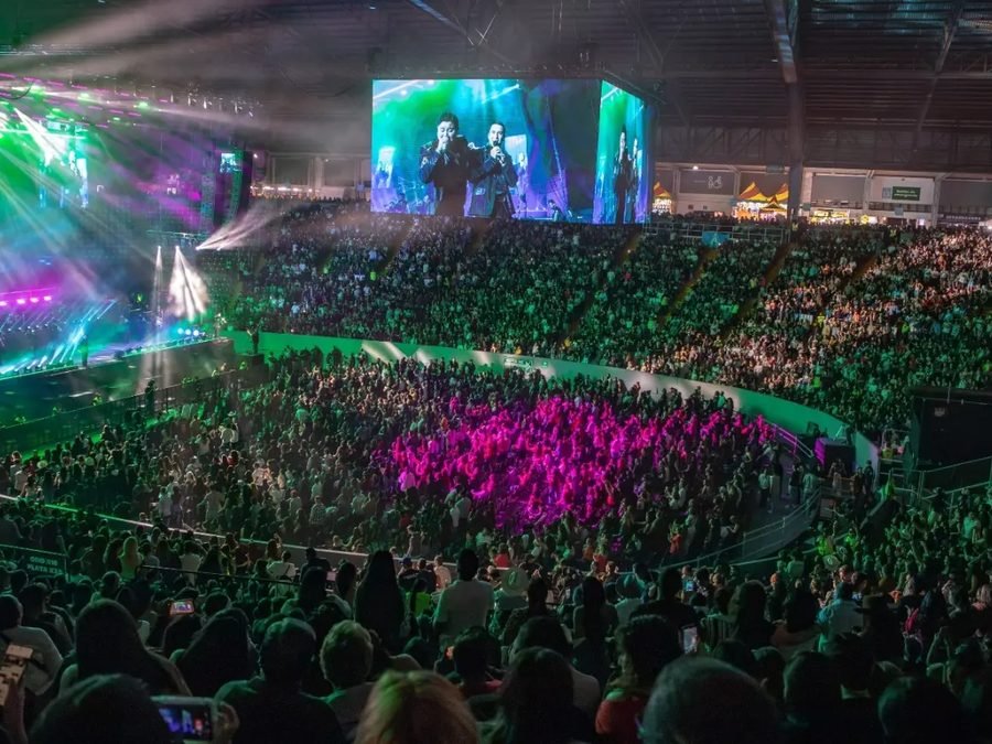 Vista panorámica del Auditorio Jalisco: pista y gradas llenas, luces verdes y moradas y las pantallas con los vocalistas