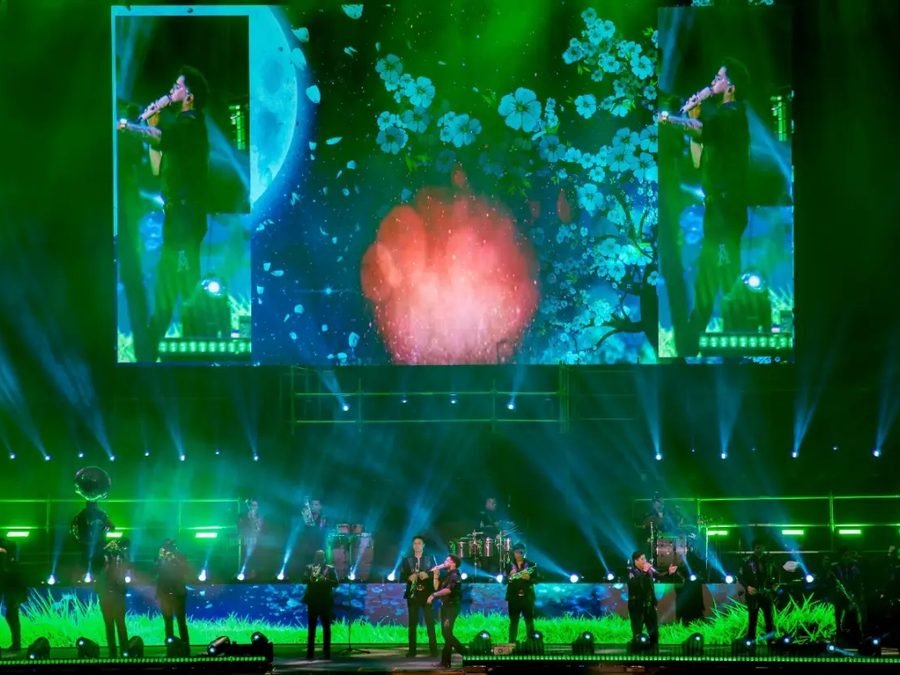 La banda alineada en el escenario bajo luces verdes; las pantallas muestran a un vocalista entre flores