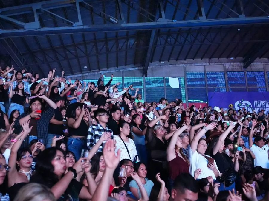 El público de las gradas canta con las manos arriba; al fondo, una lona de las Fiestas de Octubre