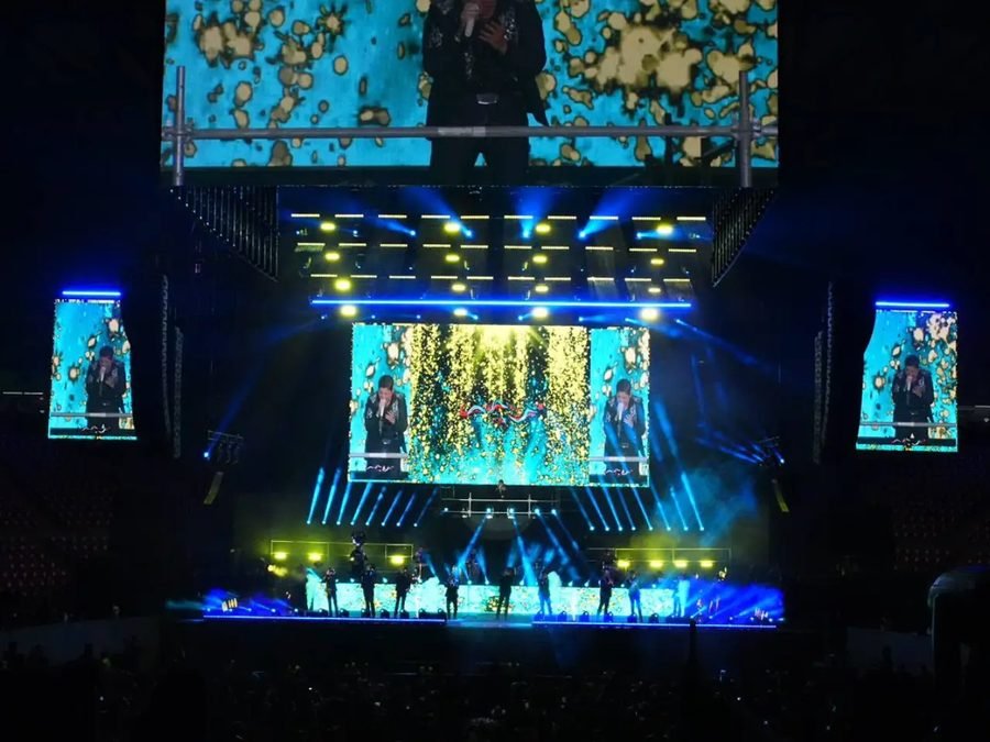 El escenario a oscuras con luces azules y amarillas; cuatro pantallas muestran a un vocalista