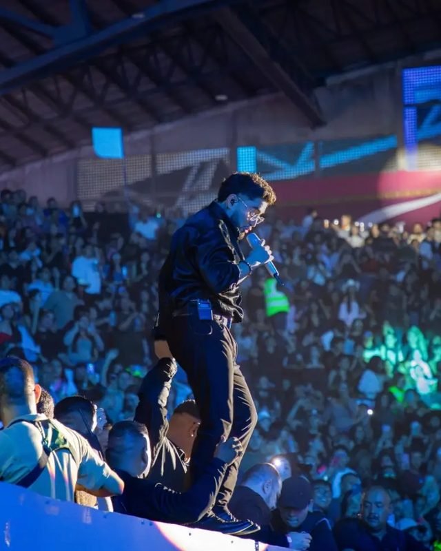 Un vocalista de chamarra negra canta de pie sobre la valla, con las gradas llenas al fondo