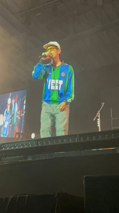 Caloncho canta al micrófono con gorra blanca, lentes amarillos y una camiseta de futbol azul y verde