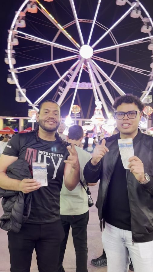 Dos amigos sostienen sus boletos afuera del Foro Principal, con la rueda de la fortuna al fondo