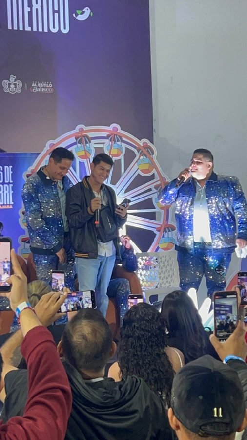 Un fan canta al micrófono entre dos integrantes de Pequeños Musical con trajes de lentejuelas en un stand de las Fiestas de Octubre, con la gente grabando