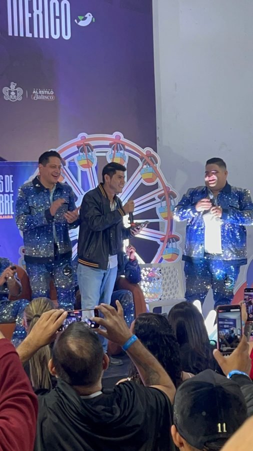 Dos integrantes de Pequeños Musical ríen junto a un fan en el stand de la feria mientras el público graba con celulares
