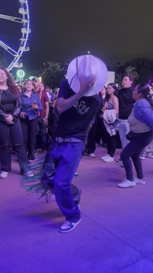 Parejas bailan en un pasillo de la feria con la rueda de la fortuna iluminada al fondo