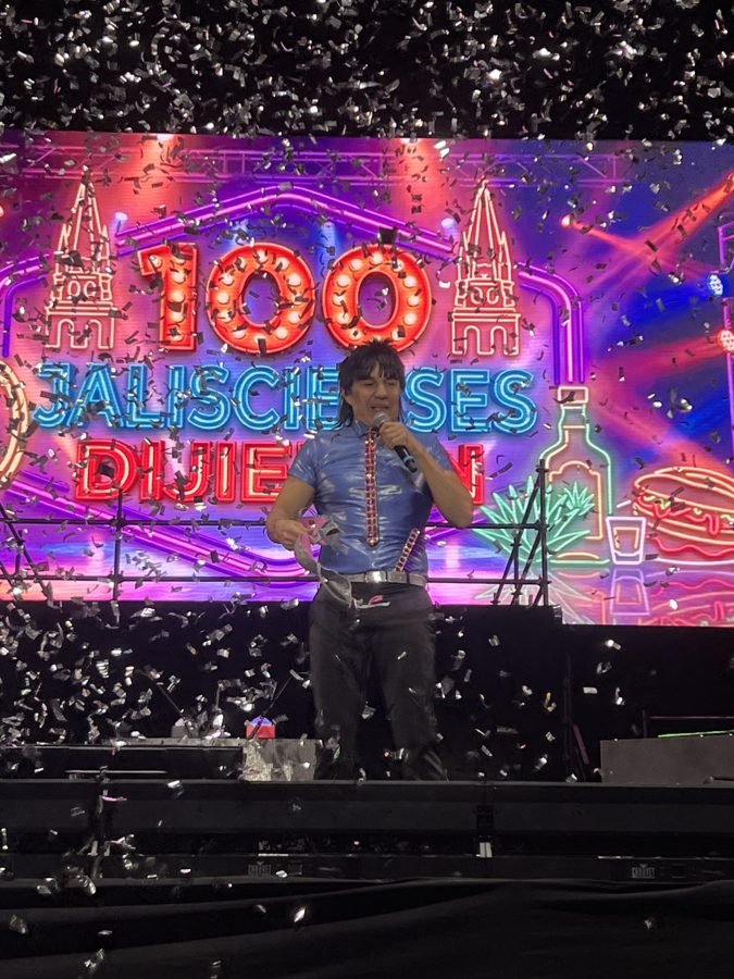 Adrián Uribe como el Vítor, con camisa azul brillante y corbata roja, bajo una lluvia de confeti plateado frente a la pantalla del show