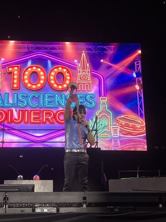 El Vítor levanta un sombrero con una mano y canta al micrófono frente a la pantalla de neón de 100 Jaliscienses Dijieron