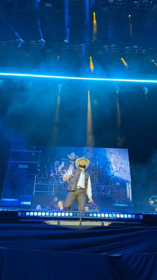 Leonardo Aguilar de sombrero blanco y chaleco bordado al frente del escenario en luz azul, con los reflectores encima