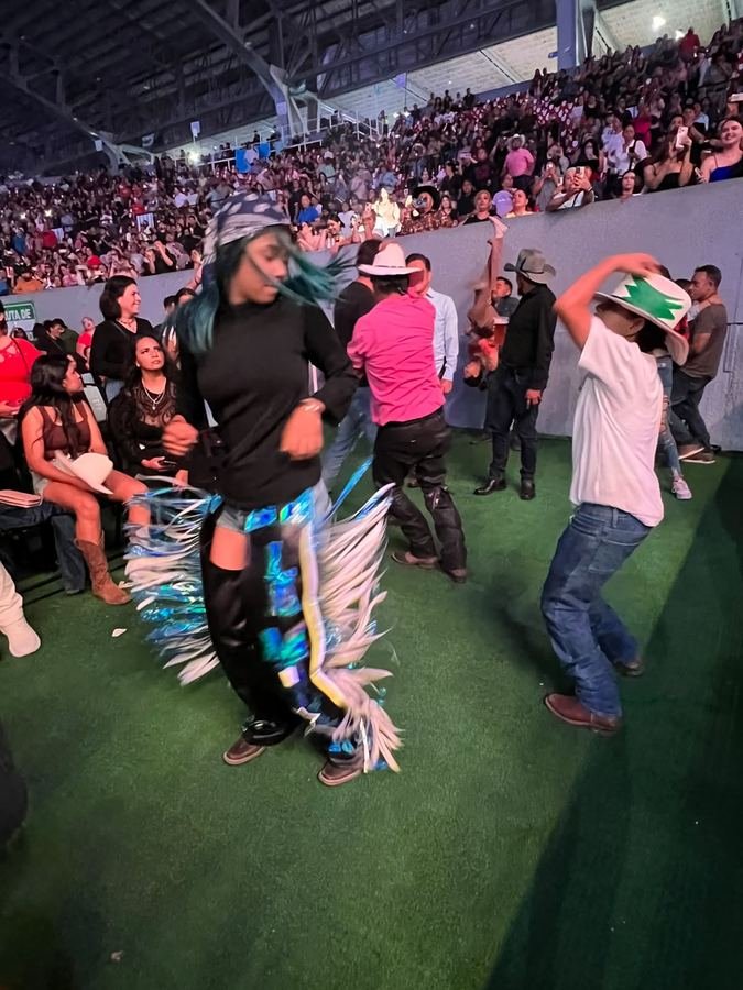 Una muchacha con chaparreras de fleco holográfico y un niño de sombrero bailan en el pasto