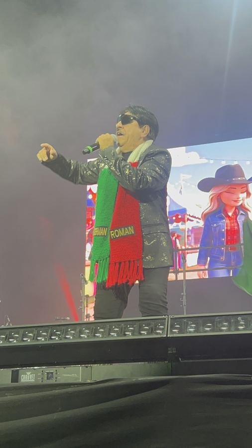 Germán Román canta con lentes oscuros, saco negro con destellos y bufanda verde, blanca y roja