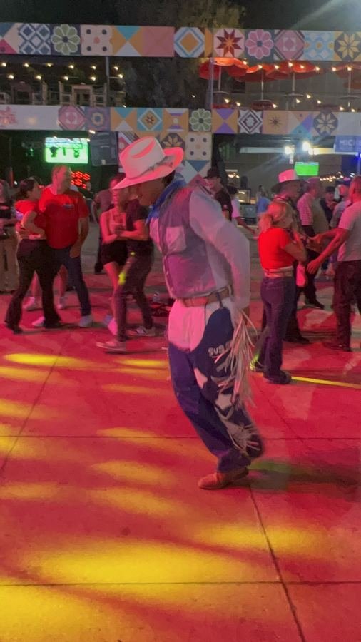 Un vaquero con sombrero blanco y chaparreras de fleco baila sobre un piso iluminado de rojo y amarillo