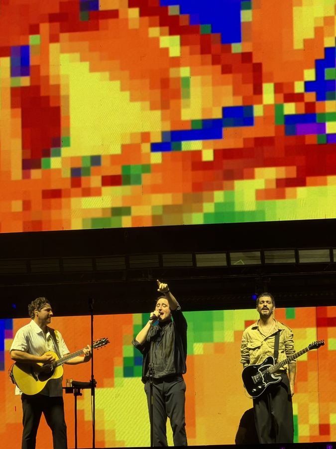 Jesús Navarro señala al público entre sus dos compañeros con guitarras frente a una pantalla de pixeles rojos y amarillos