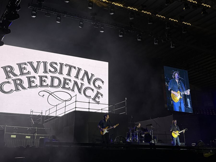 La banda completa en el escenario bajo la pantalla con el logo
