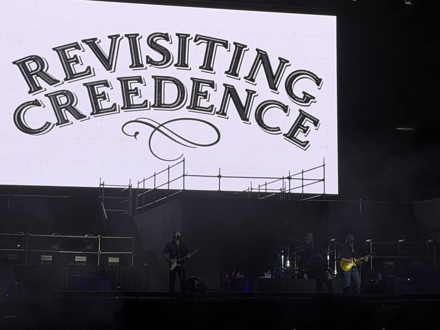La pantalla blanca con el logo de Revisiting Creedence sobre el escenario donde tocan dos guitarristas y el baterista